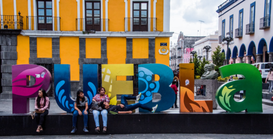 imagen en la que aparecen uno jóvenes sentados en una plaza y detrás, el nombre de la ciudad de Puebla